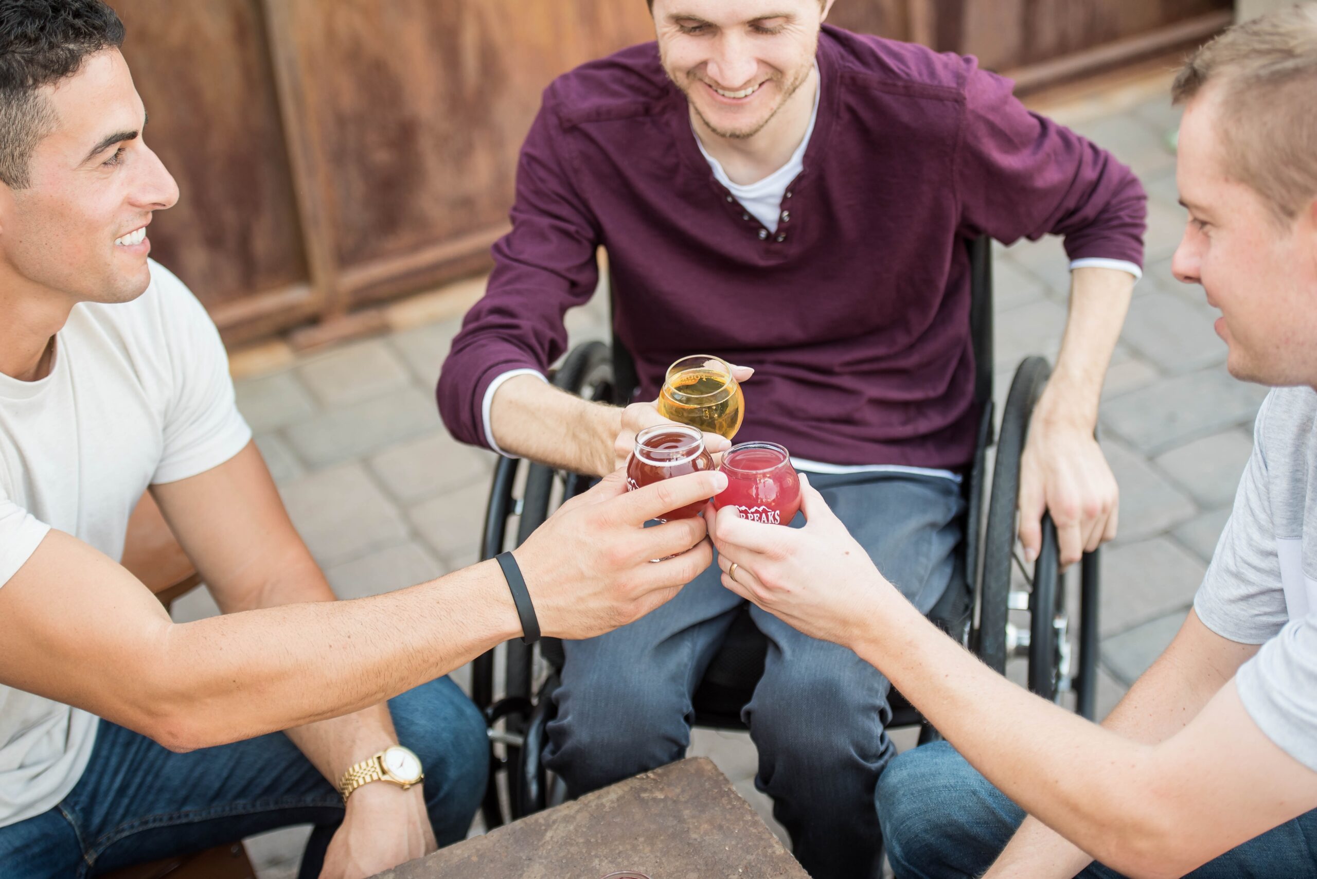 tres hombres compartiend tiempo, con bebidas en sus manos, todos sonrientes, dos de ellos sentados en sillas y uno de ellos, sentado en una silla de ruedas