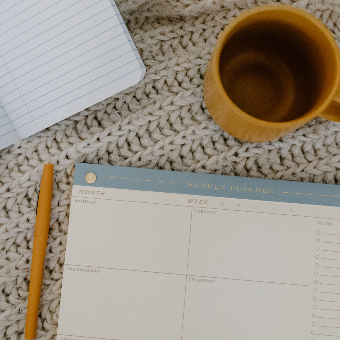 una libreta, un planificador, un bolígrafo y una taza de café, imagen que hace alusión a la planificación de la rutina de una persona