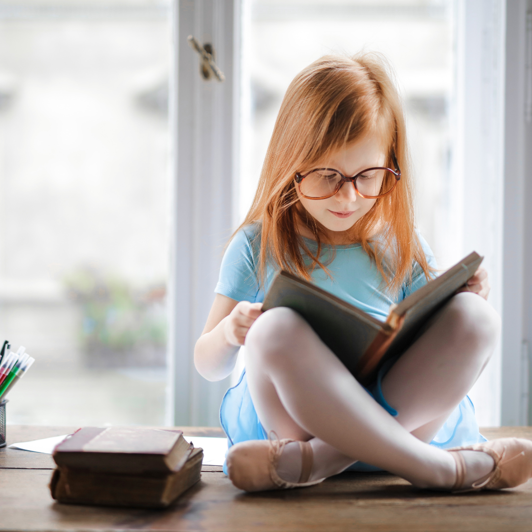 una niña sentada junto a lo que parece una ventana, con libros al lado de ella. la niña esta leyendo un libro. la imagen busca reflectar la importancia de la lectura para el cerebro